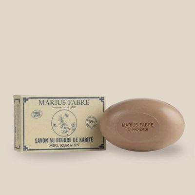 Photo produit : Savon ovale de couleur brune au beurre de karité et à l'huile d'amande douce, parfumé miel-romarin, vient dans une boîte recyclée.